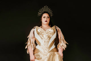 A woman wears a gold and white corset ensemble in front of a black background. The garment is embellished with feathers and she wears a gold crown headpiece