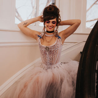 A brunette woman wears a large pale pink skirt with a set of crystal embellished stays made by the Bad Button. The woman has large hair, much like Marie Antoinette and a snake tattoo
