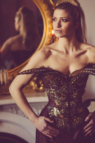 A blonde model poses in a brown corset and dress ensemble. The front of the corset has intricate bead work and long feathers extend from the top of the bust area