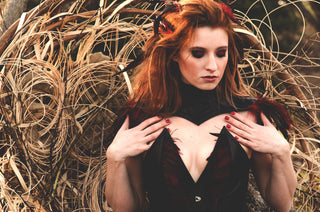 A red haired woman with red and black feathers in her hair wears a leather corset with intricate feather detailing