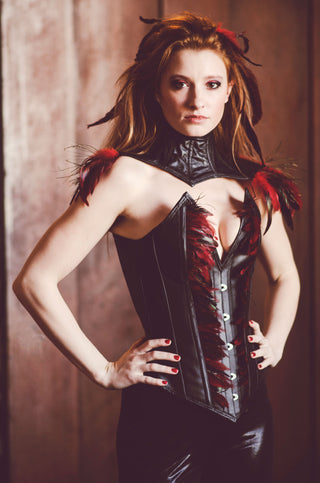 A red haired woman with red and black feathers in her hair wears a leather corset with intricate feather detailing