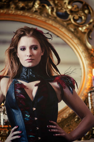 A red haired woman with red and black feathers in her hair wears a leather corset with intricate feather detailing