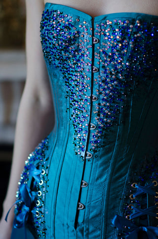 A fair red-haired model wears teal tights and a intricately beaded teal overbust corset