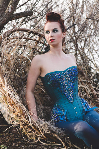 A fair red-haired model wears teal tights and a intricately beaded teal overbust corset