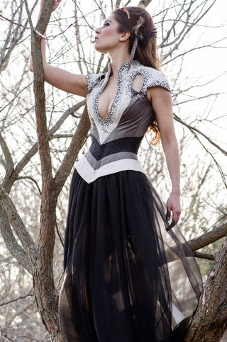 A brunette model wears a corset-ed ensemble that features grey and white panels, hand-beaded detailing and a long white tail reminiscent of a swallotail.