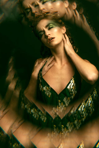 A blonde woman poses in a dark room with bold green eye make-up. She wears a green corset dress with gold scale-like embellishment