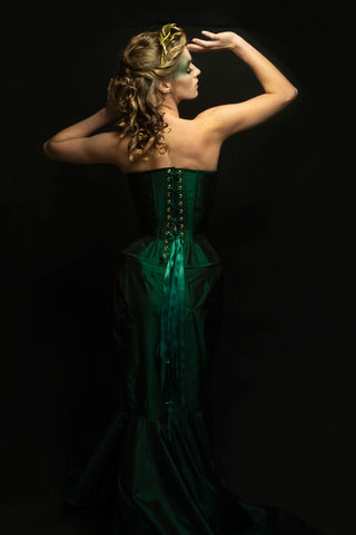 A blonde woman poses in a dark room with bold green eye make-up. She wears a green corset dress with gold scale-like embellishment