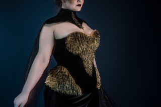 A plus sized brunette woman poses in front of a deep blue background. She wears a small gold crown and a black neck piece with a heavily embellished gold and black corset.