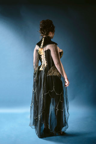 A plus sized brunette woman poses in front of a deep blue background. She wears a small gold crown and a black neck piece with a heavily embellished gold and black corset.