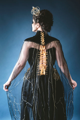 A plus sized brunette woman poses in front of a deep blue background. She wears a small gold crown and a black neck piece with a heavily embellished gold and black corset.