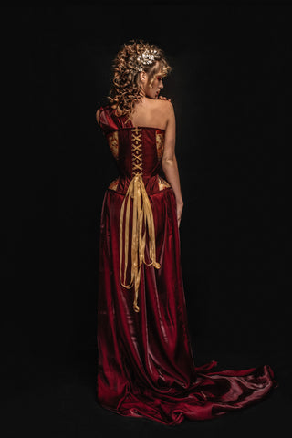 A blonde woman wears a red dress with a gold and red corset detailing. She holds a pomegranate, playing the role of persphephone