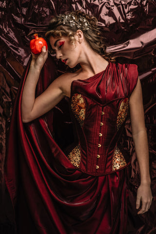 A blonde woman wears a red dress with a gold and red corset detailing. She holds a pomegranate, playing the role of persphephone