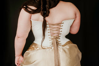 A plus sized brunette woman wears a beautiful white and gold corset dress and a gold crown in a dark black background