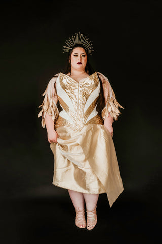 A plus sized brunette woman wears a beautiful white and gold corset dress and a gold crown in a dark black background