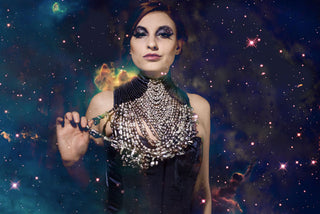 A woman in a navy embellished venetian mask wears a deep blue corset-ed dress with a high neckline with detailed pearl neck-piece. The background is galaxy themed.