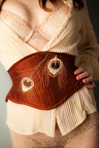 A red haired model wears a loose cream shirt with a rust coloured waspie. The mini corset features peacock feathers as an embroidered motif