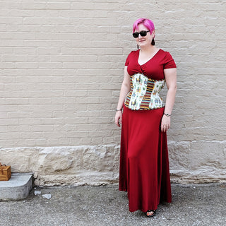 Alisha Martin, founder of The Bad Button wears a red dress and a cream coloured corset. She has pink hair and stands in front of a cream coloured brick wall