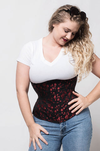 A woman in jeans and a white tee wears a red and black underbust corset by The Bad Button