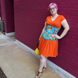 The Bad Button's founder Alisha leans against a deep pink wall while wearing a bright orange dress with bright blue patterned corset