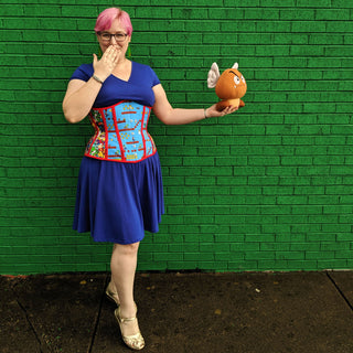 The Bad Button's founder Alisha stands in front of a green wall. She is wearing a blue dress and a novelty printed corset with red detailing