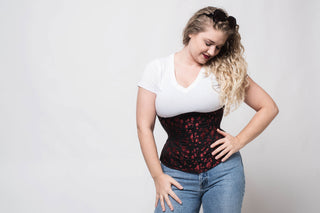 A woman with blonde hair poses in a white tee shirt and jeans. She is also wearing a The Bad Button corset that is black with red floral detailing
