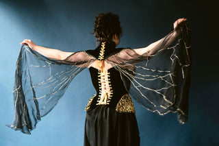 A woman wearing a gold and black corset with yellow lacing faces away from the camera. She holds her arms extended to show off delicate wings that are apart of her costume