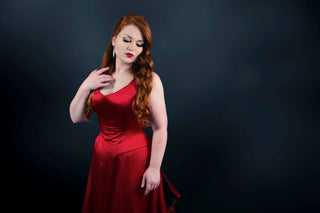 A red haired woman in a red corseted gown stands in a dark room.
