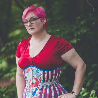 Alisha Martin stands in front of a green tree background. She has pink hair and is wearing a red shirt and a colourful blue/pink corset