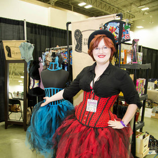 The Bad Button's Alisha stands in front of a market stall. She's wearing a bright red corset and skirt.