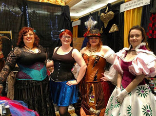 A group of four women in The Bad Button corsets stand proudly in front of a market stall. They are all dressed and accessorised in a fun, colorful way