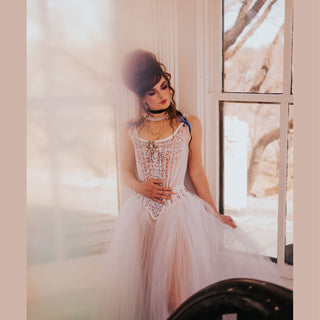 A woman with Marie Antoinette style hair is dressed in embelished stays and a light tulle skirt. She stands in a window on a stairwell. 