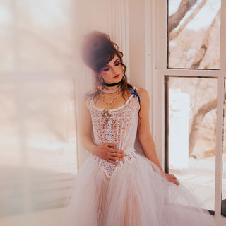 A woman with Marie Antoinette style hair is dressed in embelished stays and a light tulle skirt. She stands in a window on a stairwell. 