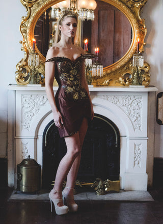 A blonde model poses in a brown corset and dress ensemble. The front of the corset has intricate bead work and long feathers extend from the top of the bust area