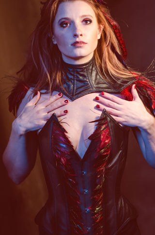 A red haired woman with red and black feathers in her hair wears a leather corset with intricate feather detailing