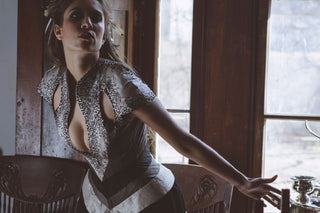 A brunette model wears a corset-ed ensemble that features grey and white panels, hand-beaded detailing and a long white tail reminiscent of a swallotail.