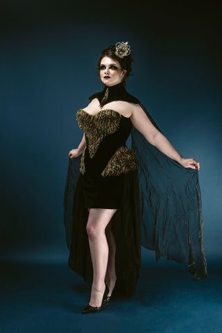A plus sized brunette woman poses in front of a deep blue background. She wears a small gold crown and a black neck piece with a heavily embellished gold and black corset. 