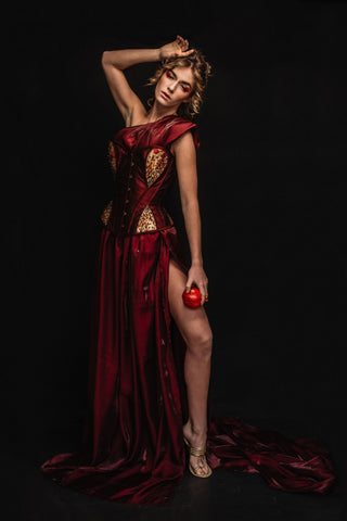 A blonde woman wears a red dress with a gold and red corset detailing. She holds a pomegranate, playing the role of persphephone