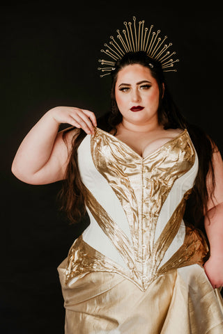 A plus sized brunette woman wears a beautiful white and gold corset dress and a gold crown in a dark black background