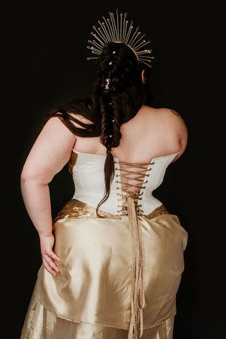 A plus sized brunette woman wears a beautiful white and gold corset dress and a gold crown in a dark black background