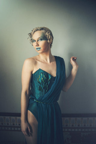 A blonde woman with bold blue makeup wears a teal corset style dress embellished with beetle shells.