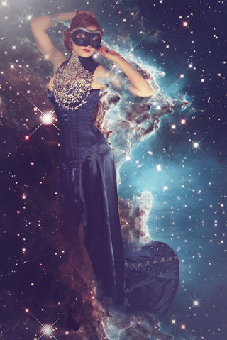 A woman in a navy embellished venetian mask wears a deep blue corset-ed dress with a high neckline with detailed pearl neck-piece. The background is galaxy themed.