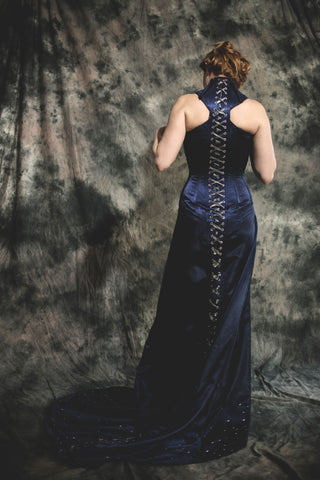 A woman in a navy embellished venetian mask wears a deep blue corset-ed dress with a high neckline with detailed pearl neck-piece. The background is galaxy themed.