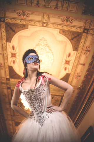 Embellished white sheer set of stays and gown with pink lacing appear on a model with a glittery venetian-style mask