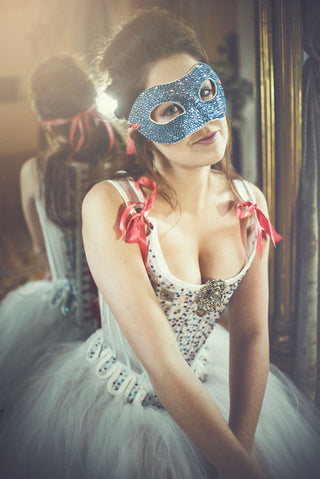 Embellished white sheer set of stays and gown with pink lacing appear on a model with a glittery venetian-style mask