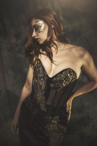 A woman stands in a grey setting with a black corseted gown with ornate gold detailing. She has gold hands, styled like Midas.