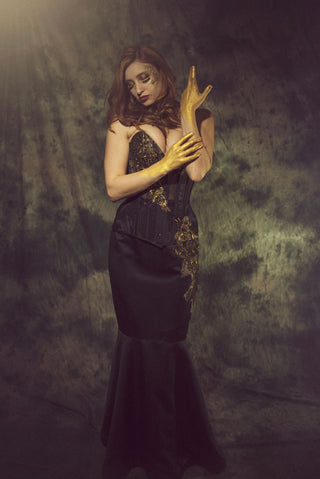 A woman stands in a grey setting with a black corseted gown with ornate gold detailing. She has gold hands, styled like Midas.