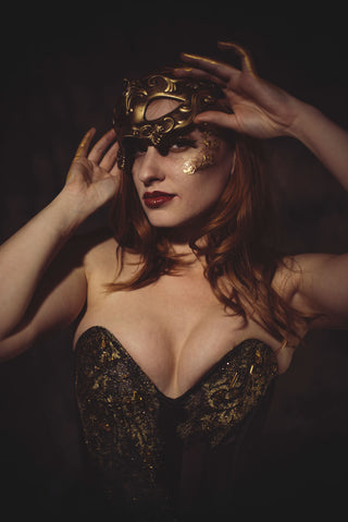A woman stands in a grey setting with a black corseted gown with ornate gold detailing. She has gold hands, styled like Midas.