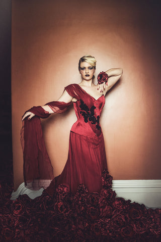 A blonde woman in a red mask stands in an ornate parlour. She wears a deep red corset dress with detailed black beading and floral elements.