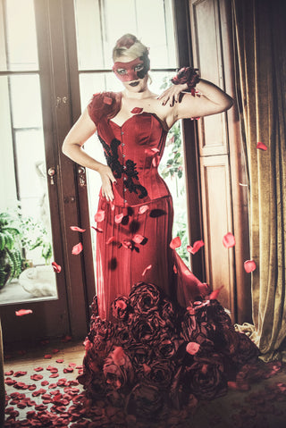 A blonde woman in a red mask stands in an ornate parlour. She wears a deep red corset dress with detailed black beading and floral elements.