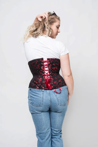 Red Rosebud Coutil black corset features delicate red floral detail. A woman in a white tee and jeans wheres the corset